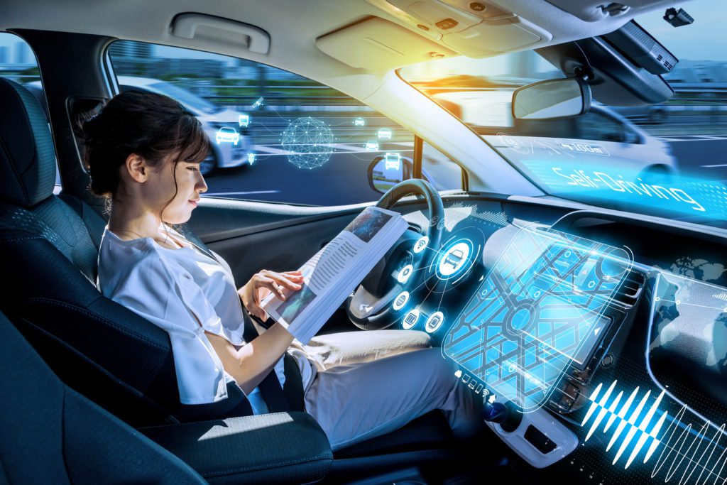 A girl sitting in the driving seat of a car reading a book while the car is driving autonomously